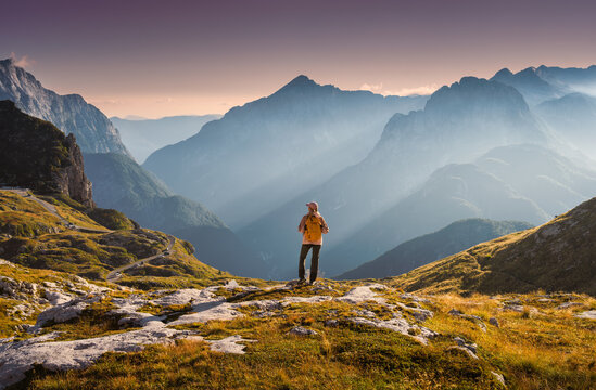 Woman hiker with backpack standing on rocky summit in autumn in Slovenia. Alpine landscape with sunbeams through fog, rocky mountain peaks, sky, clouds, orange grass, golden sunset light in fall