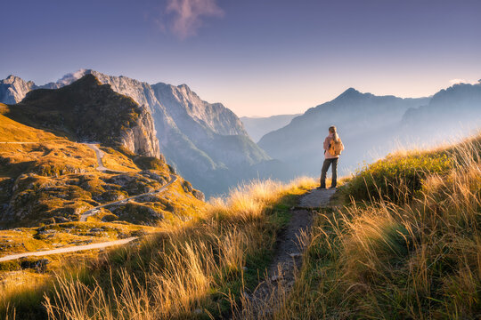 Woman hiker with backpack standing on trail at sunset. Alps in Slovenia. Alpine autumn landscape with mountain peaks, sky, clouds, golden grass, pathway, high hills, beautiful evening lighting in fall