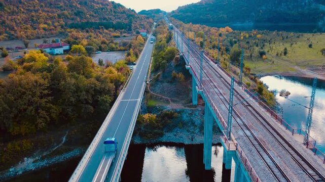 Aerial drone view of parallel railway and highway bridges over river with moving truck and passenger train in autumn.
