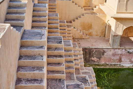 Ancient stepwell featuring symmetrical stone staircases descending in geometric patterns toward a green water reservoir. The structure showcases traditional Rajasthani architecture with weathered