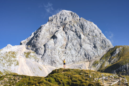Woman hiker with backpack standing on trail, looking at majestic Mangart mountain in Slovenia. Alpine autumn landscape with girl, huge rocky peak, blue sky, clouds, green grass and sunny day. Hiking