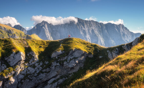 Two women hikers walking along a grassy mountain ridge, rocky peaks in clouds in the Julian Alps, Slovenia, with warm evening sunlight illuminating the alpine landscape at sunset in autumn. Nature