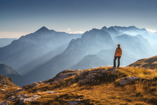 Woman hiker with backpack standing on rocky summit in autumn in Slovenia. Alpine landscape with sunbeams through fog, rocky mountain peaks, sky, clouds, orange grass, golden sunset light in fall