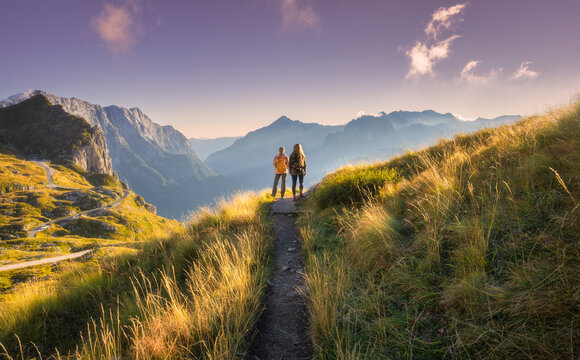 Two women hikers with backpacks standing on trail in mountain valley at sunset. Alps in Slovenia. Alpine autumn landscape with girls, mountain peaks, purple sky, clouds, golden grass, pathway in fall