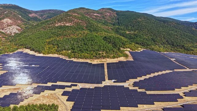 Large solar panel farm is located in a hilly region with numerous panels aligned in rows. The environment is sunny with green hills in the background.