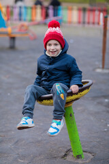 Happy little boy in a red pom pom hat and navy jacket spinning on a round playground seat in cold weather, colorful fence behind, active winter childhood concept, film grain, high quality photo © Татьяна Швец