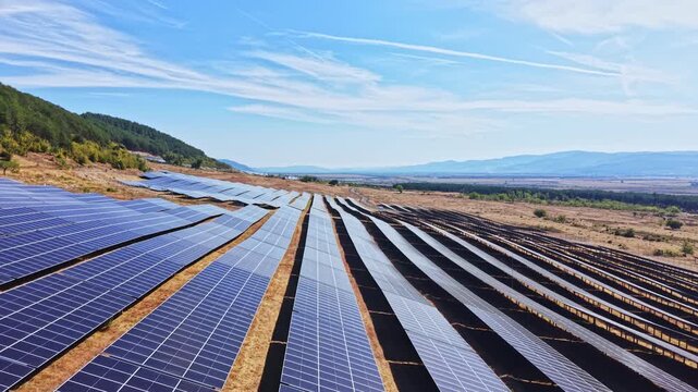 Rows of solar panels sit on the ground in a renewable energy field. The location has hills and mountains in the background. The sky is clear and sunny with some clouds.