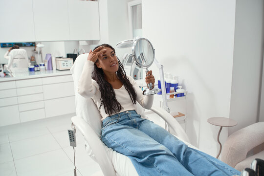 Woman checks her forehead and complexion in a handheld mirror at a beauty clinic before a skincare consultation. Acne and clogged pore concerns during facial skin self examination.