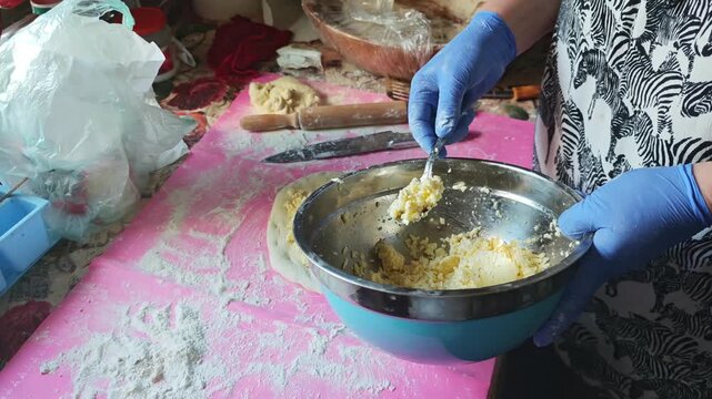 Professional baker wearing blue gloves meticulously stuffing fresh dough with a cheese filling on a pink silicone mat, preparing traditional savory pastries in a messy home kitchen environment