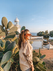 Fototapeta premium Young woman smiling beside large prickly pear cactus at coastal viewpoint with lighthouse and sea in warm golden hour light, scenic summer travel portrait by Mediterranean shore