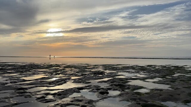 Serene sunset over rocky tidal beach with reflections and two distant silhouettes walking, Atlantic coast Morocco, 4K