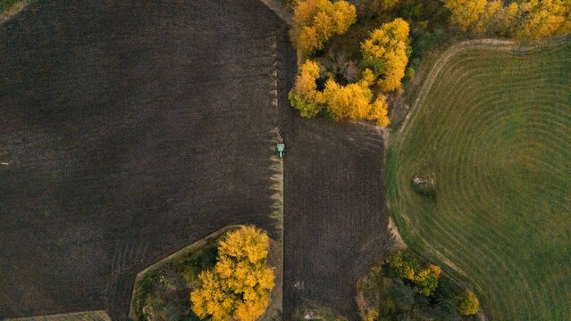 Aerial view of a tractor plowing a dark field bordered by vibrant autumn trees and green agricultural land in Finland.