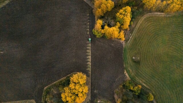 Aerial view of a tractor plowing a dark soil field bordered by vibrant yellow autumn trees and green agricultural land in Finland.