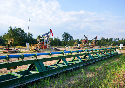 Green metal pipeline with blue pipes running across grassy field toward oil pumpjacks under partly cloudy sky