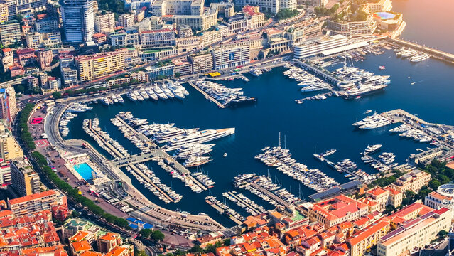 Aerial view of Port Hercules filled with luxury yachts and boats, surrounded by dense urban architecture, historic buildings, and a swimming pool along the waterfront, Monaco.