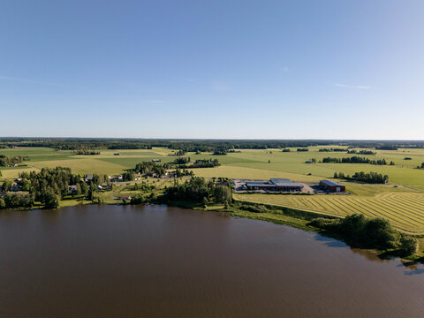 Aerial view of a serene lake bordered by lush green fields, agricultural land, and scattered farm buildings under a clear blue sky in Uusimaa, Finland.