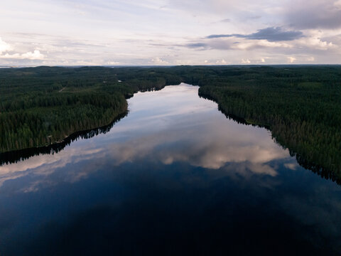 Aerial view of a serene lake surrounded by dense coniferous forests in Eastern Finland, with soft cloud reflections on the calm water surface, Finland.