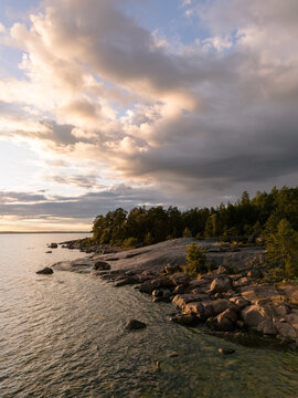 Aerial view of the rocky coastline of Southern Finland with dense forest, calm water, and large boulders under a dramatic sunset sky in Finland.