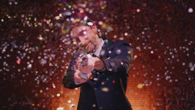 Attractive young man in suit blows up a popper at a party, having fun. Handsome male explosion a firecracker in his hands. Celebrating or holiday confetti. Victory at festival, congratulations.