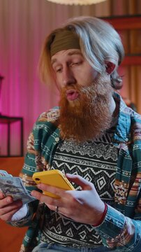 Planning family budget. Bearded redhead man counting money dollar cash use smartphone calculate domestic bills at home. Joyful hippie guy satisfied of income saves money for planned vacation, gifts