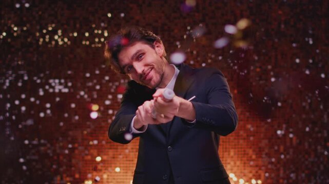 Attractive young man in suit blows up a popper at a party, having fun. Handsome male explosion a firecracker in his hands. Celebrating or holiday confetti. Victory at festival, congratulations.