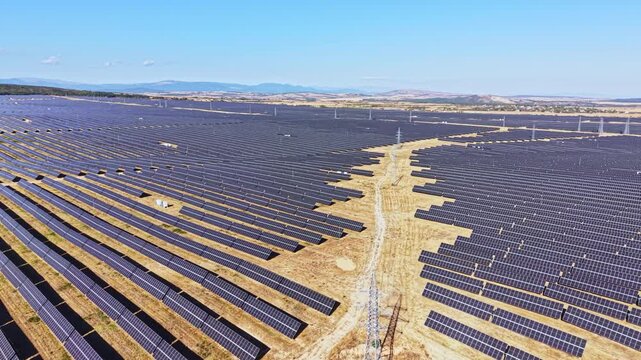 Rows of solar panels fill the landscape in a large solar farm. The sun is shining, and there are no clouds in the sky. The area is open with some mountains in the background.