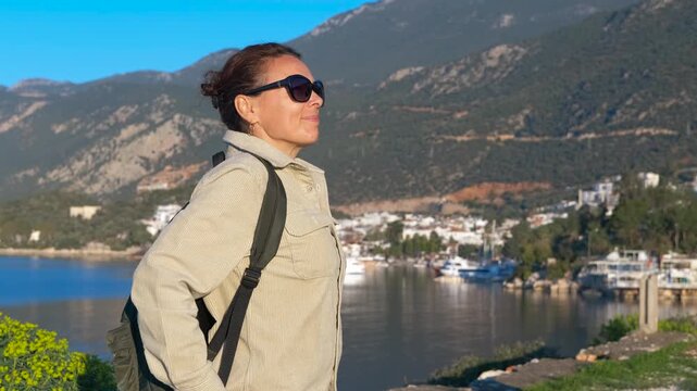 Tourist enjoying kas marina view in antalya, turkey. Female traveler wearing sunglasses and backpack standing on viewpoint, gazing at panoramic seascape of historic kas marina