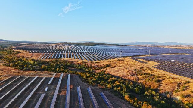 Large solar panel installation covers the ground in a rural landscape. Green patches and distant mountains are visible under clear blue skies during late afternoon hours.