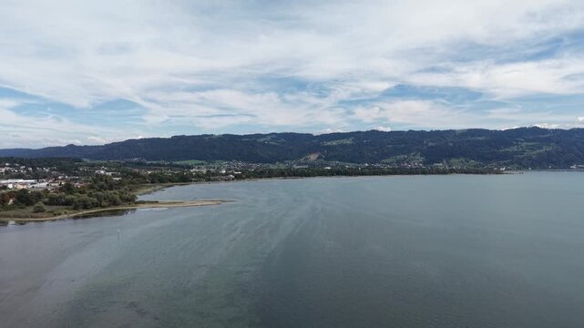 Aerial panoramic view of the eastern shore of Lake Constance near Bregenz, Austria, showing shallow turquoise water, exposed sandbanks, green wetlands, lakeside settlements and forested hills beneath 