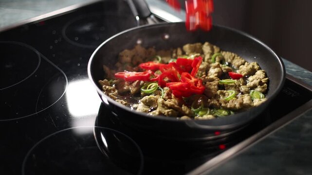 Red peppers fall into a pan with leeks and pieces of soy on a stove