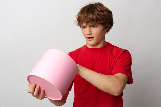 Young man in red t-shirt holds a pink container and gives a suspicious side-eye glance, expressing doubt and distrust while checking a questionable package on gray background