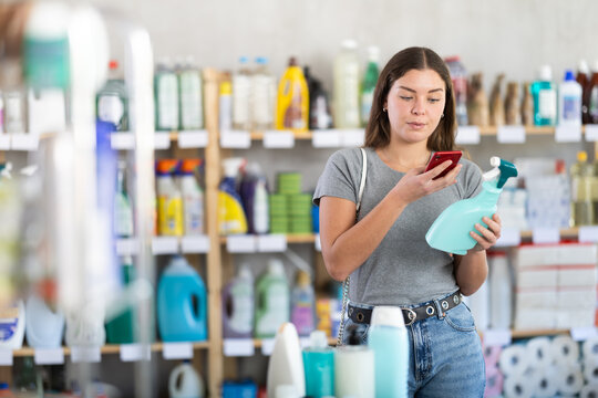 Interested smiling young female customer using mobile app to scan barcode on disinfectant spray for discounts and promotional offers during shopping in cleaning supply store..