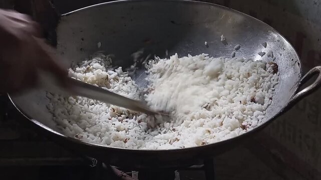 Rice cooks in a large metal wok. Hands stir grains with a wooden spoon. Steam rises from hot, fluffy rice. The wok sits on a simple stove burner. Warmth and comfort radiate from the scene