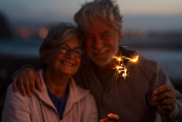 Senior couple embraces while celebrating love with glowing sparklers outdoors at dusk. Smiling elderly partners share a joyful retirement anniversary moment, illuminated by warm sparks and twilight. © Daniel