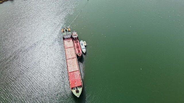 A cargo ship is anchored in green water with a smaller vessel nearby. The scene shows a harbor area during the day with some floating objects in the water.