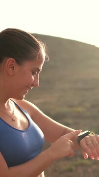 Woman checks her smartwatch and taps the screen after an outdoor fitness workout. She smiles in golden sunset light in a hilly landscape while monitoring exercise stats, health and active lifestyle.