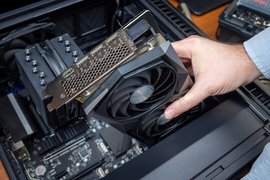 A technician installs a powerful triple-fan gaming graphics card on a gaming PC motherboard. A dual-tower CPU cooler and black power cables are visible