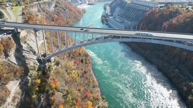 Aerial over the Niagara River, international border crossing Queenston-Lewiston bridge between New York and Canada, semi trucks with Niagara Falls hydroelectric plants. Autumn fall foliage trees.