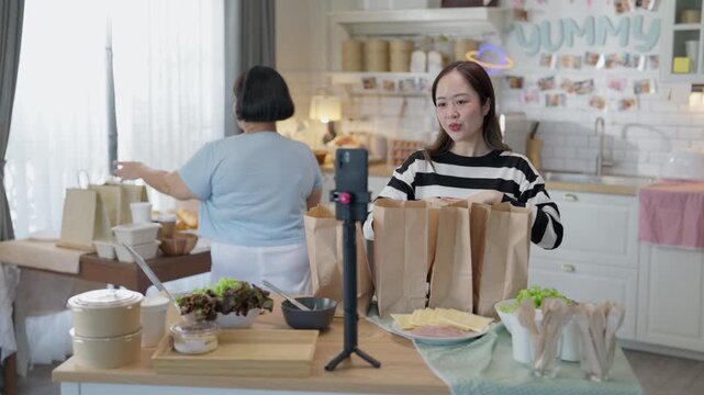 Cooking Baking Kitchen Live Streaming Online with Two Asian Women Presenting Homemade Packaged Food and Paper Bags During Cheerful Adult Home Culinary Broadcast for Viewers