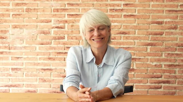 Senior woman smiles at camera, looks away thoughtfully, then rests her face in her hands and smiles again at a home office table. Static shot conveys reflection, aging, work-life balance and health.