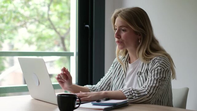 Woman works on a laptop at her home desk, then pauses typing and looks thoughtfully out the window. Calm remote work with coffee, notebook and daylight conveys focus, reflection and contemplation.