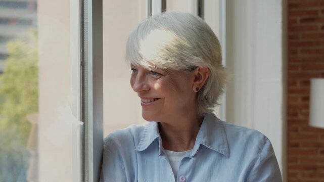 Senior woman stands by a sunlit window in her home office, smiling and looking outside. She pauses from remote work to reflect, showing calm, wellbeing, aging, and work-life balance.