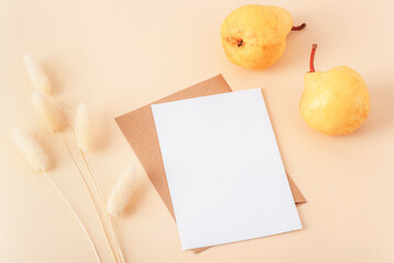Two golden pears beside a blank white card and kraft envelope on a beige background with bunny tail grass. Top view, mockup © Viktoriya