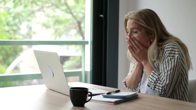 Woman working on a laptop at home reacts with shock, then celebrates good news by raising her fists, laughing and clapping. The joyful remote worker sits at a desk, looking at her computer screen.