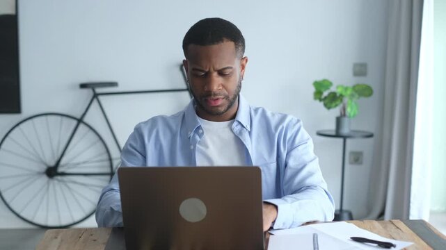 Man works on a laptop at his home office desk, then stops typing and rubs his temples in stress. Frustrated remote worker shows headache, fatigue, burnout and pressure during a difficult work task.