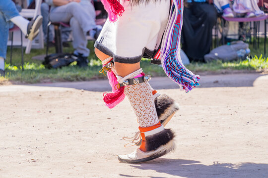 Detailed view of colorful fringed skirt, beaded garters, and moccasins during a cultural performance on a dirt ground.