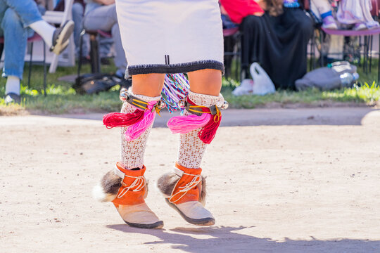 Detailed view of colorful fringed skirt, beaded garters, and moccasins during a cultural performance on a dirt ground.