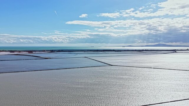 Large salt flats are visible near the ocean. The blue water and a clear sky with clouds add to the landscape. The scene shows rows in the salt flats under natural light.