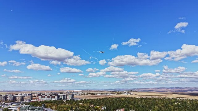 Airplane flies through a blue sky filled with clouds above a city with buildings in urban settings. The scene shows land and hills in distance.