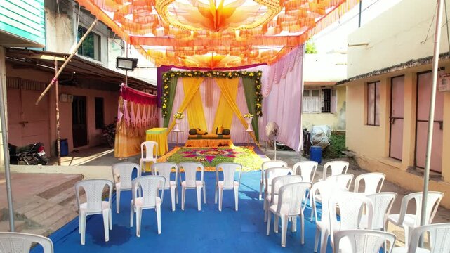 The camera moves back from a courtyard ceremony setup with orange overhead fabric, a yellow floral backdrop, rows of white chairs and a blue carpet.